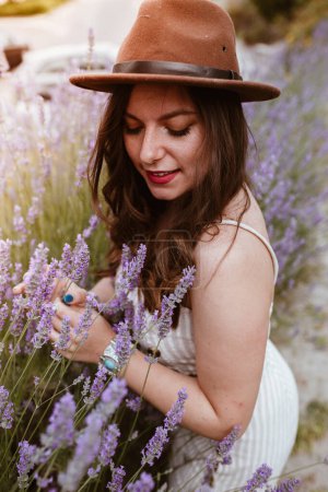 Photo for Attractive long haired woman in hat enjoying lavender and smiling with eyes closed - Royalty Free Image