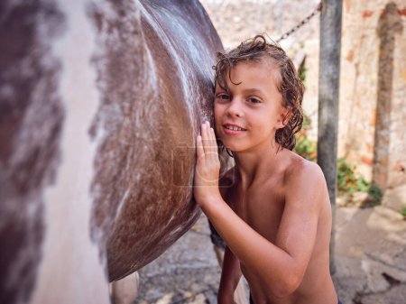 Smiling shirtless boy with wet hair touching horse body while looking at camera near old brick wall in countryside in summer