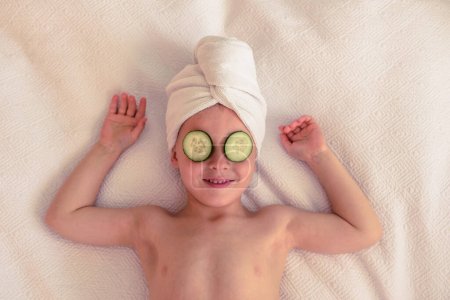 Téléchargez les photos : Du haut du petit enfant souriant avec une serviette sur la tête et des tranches de concombre sur les yeux reposant sur une couverture blanche avec les bras levés - en image libre de droit