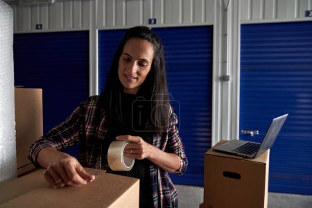 Téléchargez les photos : Femme positive dans des vêtements décontractés emballant des biens dans une boîte avec du scotch tout en se tenant près d'un ordinateur portable et d'un self-stockage fermé - en image libre de droit