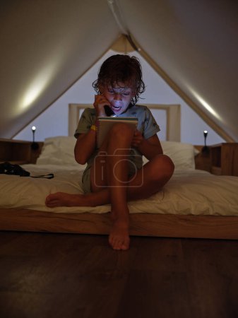 Photo for Full body of amazed barefoot boy reading interesting book with flashlight while sitting on bed in mansard with dim light - Royalty Free Image