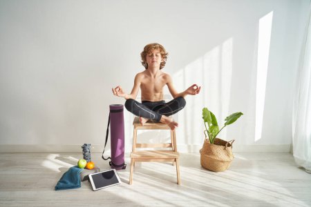 Full body of barefoot preteen boy sitting with crossed legs on wooden stool and relaxing in Padmasana yoga pose