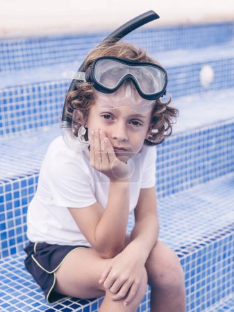 Foto de Lindo niño preadolescente en máscara de snorkel apoyado cabeza en la mano mientras está sentado en los escalones de baldosas de la piscina y mirando a la cámara - Imagen libre de derechos