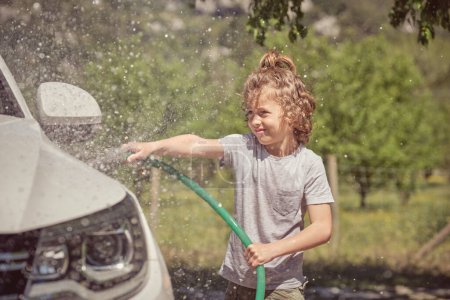 Téléchargez les photos : Petit enfant éclaboussant des gouttes d'eau tout en lavant la voiture avec tuyau par une journée ensoleillée d'été sur fond flou - en image libre de droit