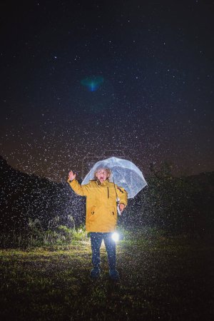 Foto de Cuerpo completo de niño anónimo en traje amarillo de pie con paraguas y brazo levantado bajo el oscuro cielo nocturno durante la lluvia - Imagen libre de derechos