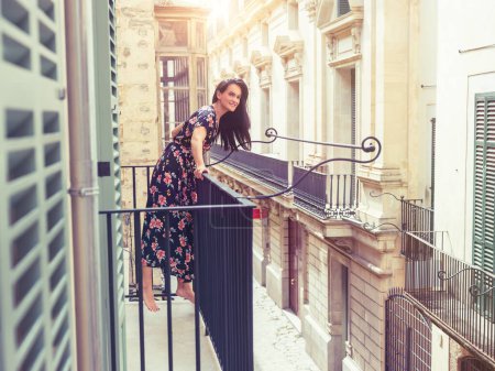 Téléchargez les photos : Vue latérale de la jeune femme souriante pieds nus avec de longs cheveux foncés portant une robe longue avec une impression de fleurs debout sur le balcon et appuyée sur une clôture en métal tout en regardant loin - en image libre de droit