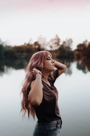 Téléchargez les photos : Vue latérale de belle femme avec de longs cheveux roses debout contre un lac tranquille avec les yeux fermés - en image libre de droit