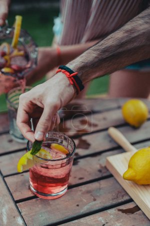 Téléchargez les photos : Cultiver homme moderne adulte garnissant cocktail glacé rose avec du citron et feuille de menthe sur dessus de table en bois - en image libre de droit