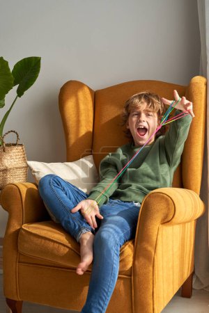 Foto de Retrato de niño riendo en ropa casual sentado en sillón y divirtiéndose mientras hace figuras de cuerdas con cuerdas de colores en casa - Imagen libre de derechos