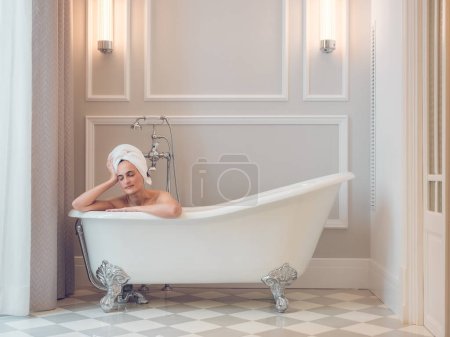 Photo for Gorgeous female with towel on head sitting in white soaker tub in light classic bathroom in daytime - Royalty Free Image
