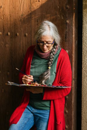 Foto de Encantada artista femenina senior con paleta de pintura y pinceles de pie cerca de la puerta de madera - Imagen libre de derechos