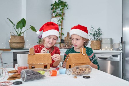 Photo for Cute siblings in red Santa hats gluing details of sweet gingerbread houses with icing in light kitchen during Christmas preparation - Royalty Free Image