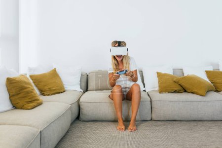 Photo for Full body of barefoot female in modern VR goggles playing videogame with gamepad while sitting on couch in living room - Royalty Free Image
