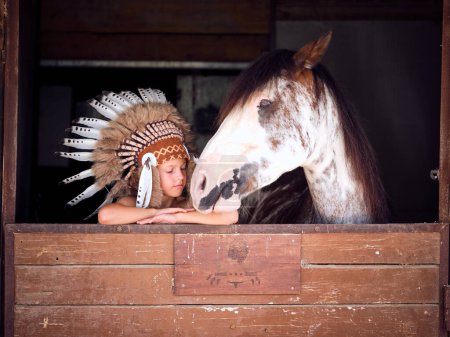 Foto de Adorable niño en traje de nativo americano apoyado en la cerca de establo cerca de blanco manchado amigo caballo - Imagen libre de derechos