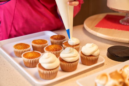 Téléchargez les photos : Personne de culture méconnaissable décoration cupcakes à l'aide de sac de tuyauterie debout au comptoir de la cuisine avec d'autres desserts sur elle - en image libre de droit