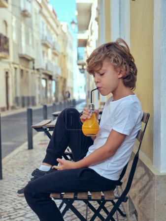 Photo for Side view of cute boy drinking fresh juice from bulb shaped bottle while sitting on street near building in city - Royalty Free Image