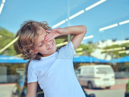 Portrait of positive child in casual clothes standing on street against window and rubbing eye while enjoying bright summer day