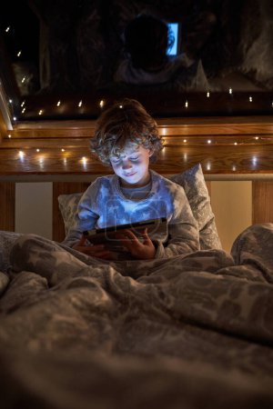Photo for Focused boy browsing tablet while lying on comfortable bed under blanket near window with glowing garland on night time in bedroom - Royalty Free Image