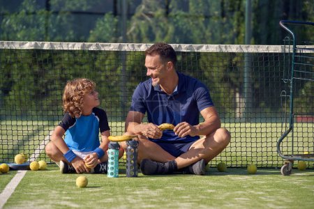 Photo for Full body of happy trainer and boy looking at each other while sitting with bananas near net during padel training - Royalty Free Image