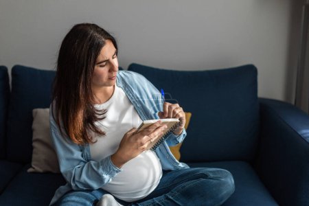 Téléchargez les photos : Femme enceinte concentrée assise sur un canapé et écrivant dans un bloc-notes tout en se refroidissant dans un salon confortable - en image libre de droit
