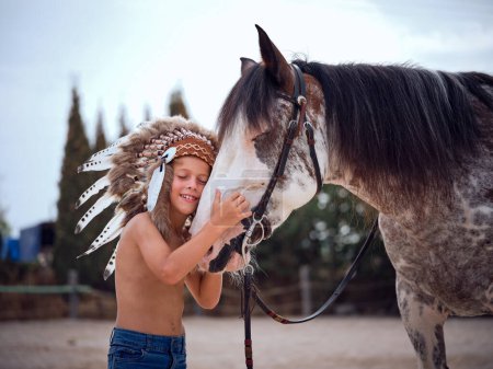 Foto de Alegre joven abrazando caballo de pie con los ojos cerrados mientras descansa cabeza a caballo en el paddock bajo la luz del sol azul - Imagen libre de derechos