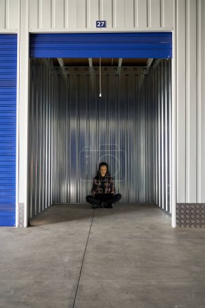 Photo for Full body of calm female in casual wear sitting in empty container of self storage with opened rolled up door during moving day - Royalty Free Image