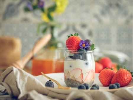 Téléchargez les photos : Délicieux yaourt aux fraises mûres et bleuets décorés de fleurs servis en pot sur la table dans la cuisine domestique - en image libre de droit