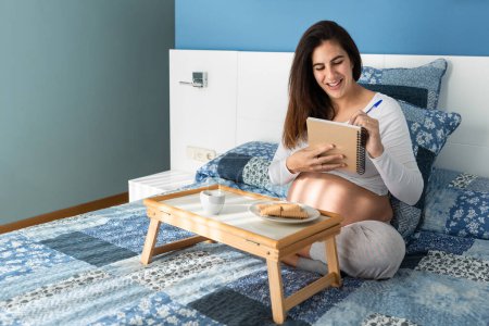 Téléchargez les photos : Femme enceinte assise au lit et écrivant dans un cahier pendant le petit déjeuner le matin - en image libre de droit