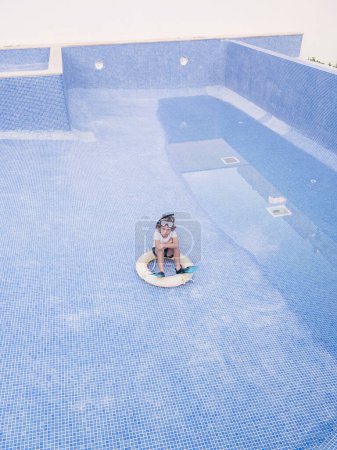 Photo for High angle of boy wearing flippers and snorkeling mask sitting on swimming ring on tiled bottom of blue pool embracing knees and looking at camera in daylight - Royalty Free Image