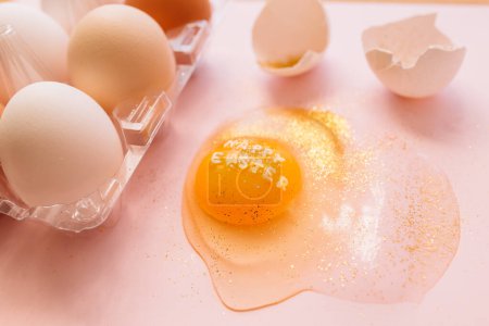 Foto de Vista superior de huevo roto brillante con texto Feliz Pascua y huevos crudos en caja de plástico sobre fondo rosa - Imagen libre de derechos