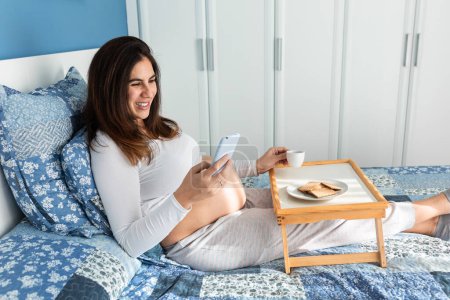 Foto de Alegre embarazada sentada en la cama por la mañana y desayunando mientras navega por el teléfono inteligente - Imagen libre de derechos