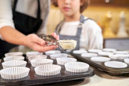 Téléchargez les photos : Cultivez une femelle méconnaissable avec une cuillère de pâte à cupcake debout près du fils dans un tablier à table avec des casseroles de cuisson dans la cuisine légère - en image libre de droit