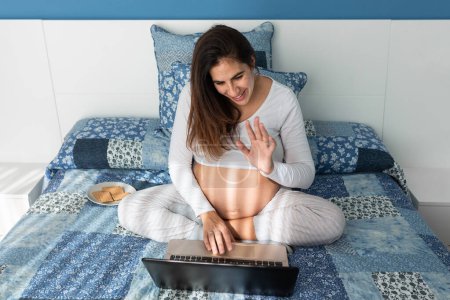 Photo for From above of smiling pregnant female speaking on video call on netbook and waving hand while sitting on comfortable bed at home - Royalty Free Image