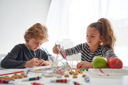 Téléchargez les photos : Enfants concentrés en tenue décontractée peignant avec des pinceaux assis à table avec des crayons éparpillés et de la nourriture dans la pièce lumineuse - en image libre de droit