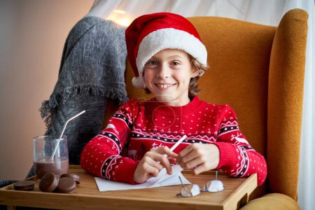 Photo for Glad kid in Santa hat painting at table with glass of chocolate and cookies during Christmas celebration at home - Royalty Free Image