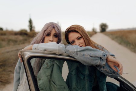Téléchargez les photos : Femmes confiantes en jean veste debout avec cigarette à la porte de la voiture regardant caméra dans le pays - en image libre de droit