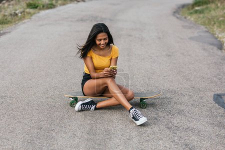 Foto de Navegación femenina étnica en el teléfono inteligente mientras está sentado longboard en el camino de asfalto - Imagen libre de derechos