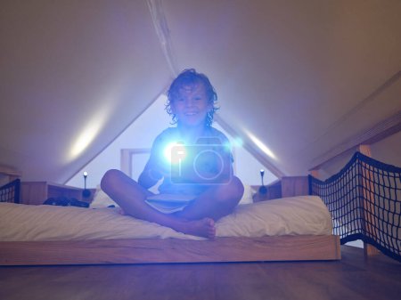 Photo for Full body of cheerful boy with notebook in casual wear shining flashlight and looking at camera while sitting on bed in mansard - Royalty Free Image