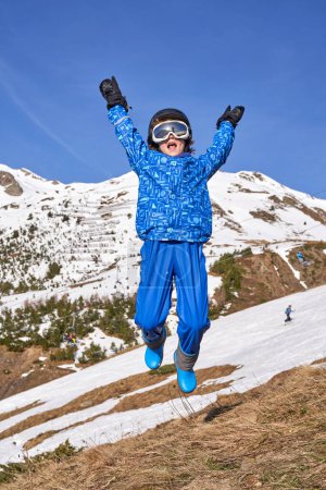 Foto de Niño feliz en ropa de abrigo con gafas y casco levantando brazos y gritando mientras salta en la pendiente contra las montañas nevadas y el cielo azul en el día de fin de semana en la estación de esquí - Imagen libre de derechos