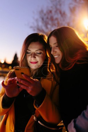 Téléchargez les photos : Contenu amies en vêtements chauds décontractés souriant tout en surfant téléphone mobile avec ciel du soir sur fond flou - en image libre de droit