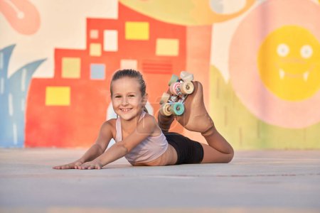 Photo for Positive little girl in roller skates lying on belly and putting hands in front of body in crop top and shorts while looking at camera during rest - Royalty Free Image