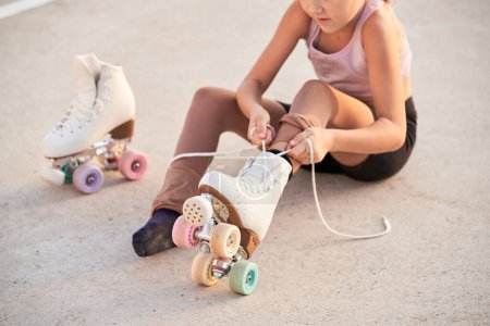 Téléchargez les photos : Crop fille anonyme attacher lacets sur patins à roulettes quad avec roues colorées tout en étant assis sur l'asphalte passerelle pendant l'entraînement sur la rue - en image libre de droit