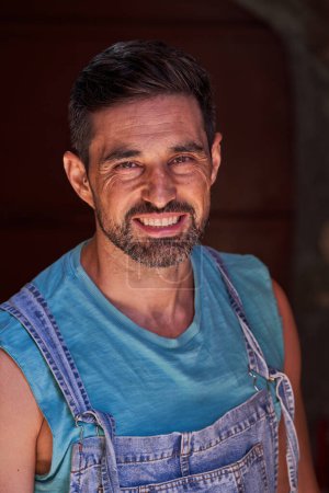 Photo for Positive worker with beard in t shirt and denim overall looking at camera with smile while standing in workshop during workday - Royalty Free Image