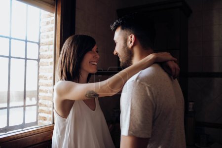 Photo for Side view of pretty woman smiling and hugging handsome man while standing near window in stylish kitchen together - Royalty Free Image