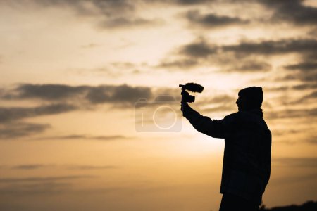 Photo for Side view silhouette of unrecognizable young female blogger in hat shooting video on digital camera against sunset sky in countryside - Royalty Free Image