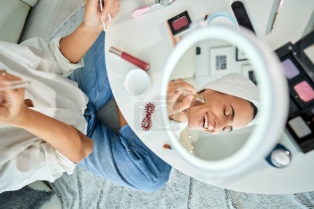 Photo for From above of reflection in mirror of positive female sitting on floor and applying moisturizing cosmetic mask on face with brush while relaxing with closed eyes during skin care procedure - Royalty Free Image