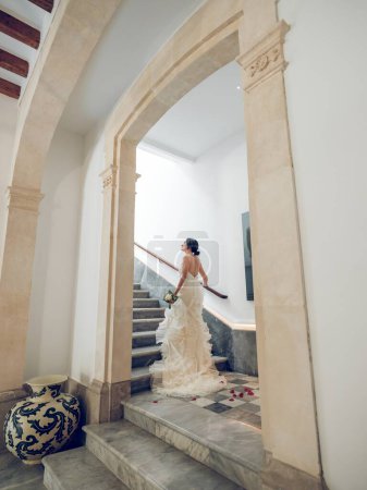 Photo for Back view of full body young female newlywed wearing elegant bridal dress and with bunch of flowers looking up while walking upstairs holding on banister in hallway of hotel - Royalty Free Image