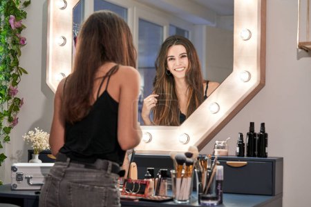 Foto de Encantada joven morena tocando el pelo mientras se mira en el espejo en el estudio de belleza y haciendo maquillaje con coloridos cosméticos decorativos - Imagen libre de derechos