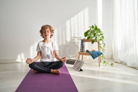 Photo for Full body of concentrated curly haired teen boy meditating in Padmasana pose while practicing yoga sitting with crossed legs on mat - Royalty Free Image