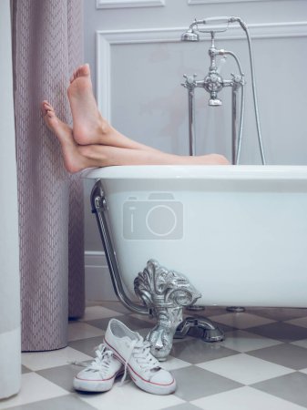 Téléchargez les photos : Femme pieds nus méconnaissable reposant dans une baignoire blanche avec les jambes hors de l'eau et des baskets dans la salle de bain légère - en image libre de droit